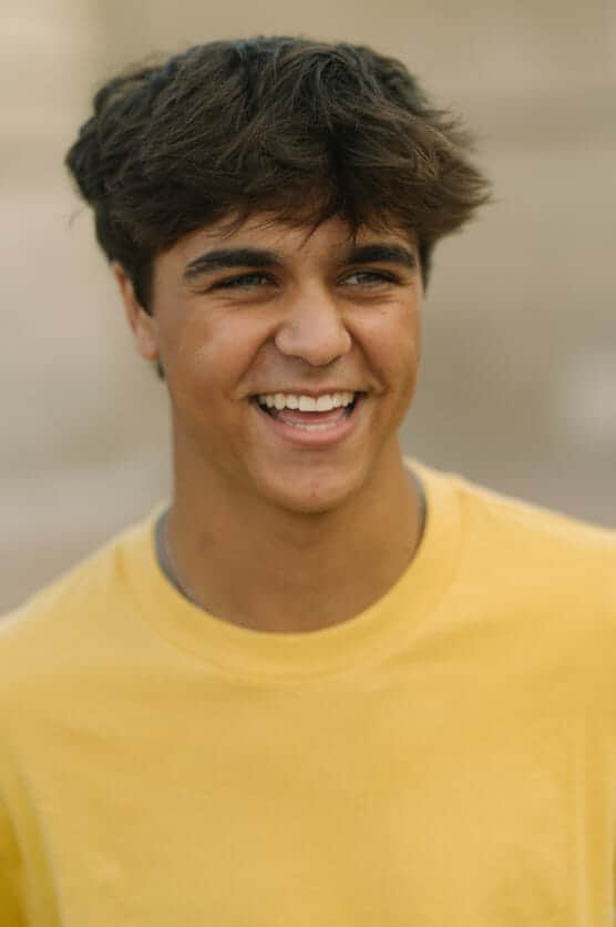 A young man with short, tousled brown hair wearing a yellow T-shirt is smiling and looking slightly to the side. The background is softly blurred, drawing focus to his joyful expression. by Justin Salem Meyer