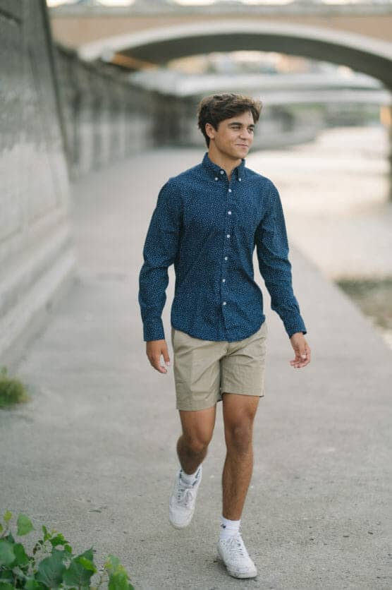 A young man wearing a blue button-up shirt, beige shorts, and white sneakers walks along a paved riverside path with bridges in the background. by Justin Salem Meyer