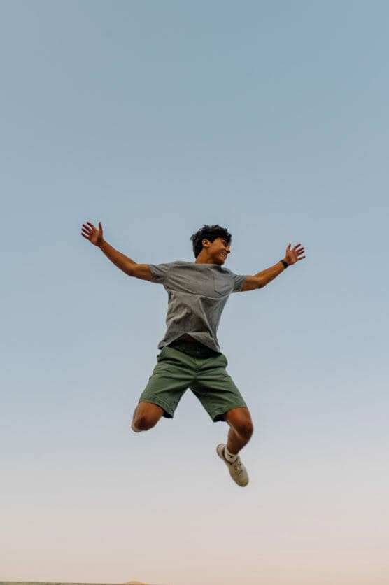 A person wearing a gray t-shirt, green shorts, and white shoes is jumping with arms outstretched against a clear blue sky. by Justin Salem Meyer