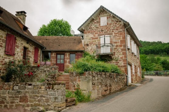 A rustic stone house with red shutters and a small balcony sits beside a quiet road, surrounded by greenery and a stone wall, with trees and hills in the background under an overcast sky. by Justin Salem Meyer