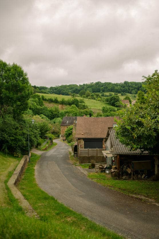 A narrow, winding rural road curves past old stone houses with tiled roofs, surrounded by lush green trees and fields under a cloudy sky. by Justin Salem Meyer