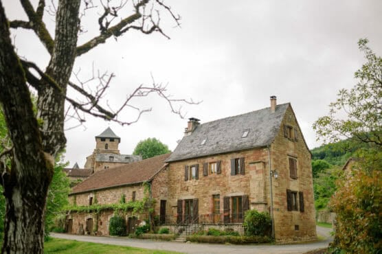 A rustic stone house with wooden shutters and a sloped roof stands beside a narrow road, surrounded by greenery and trees, with another stone building visible in the background under a cloudy sky. by Justin Salem Meyer