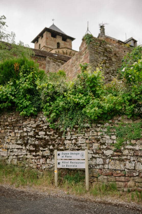 A weathered stone wall covered in green vines stands beside a road, with a directional sign pointing to “Espace Geologie St Pierre” and “Hotel Restaurant La Bastide.” An old stone building rises in the background. by Justin Salem Meyer