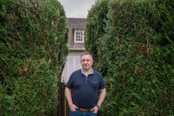 A man stands outdoors between two tall, neatly trimmed hedges, with a house in the background. He is wearing a dark polo shirt and jeans, with his hands in his pockets, smiling at the camera. by Justin Salem Meyer