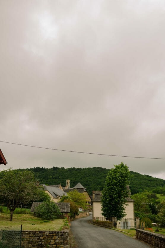 A narrow road curves past stone houses with slate roofs in a small rural village, surrounded by green trees and hills under a cloudy, overcast sky. by Justin Salem Meyer