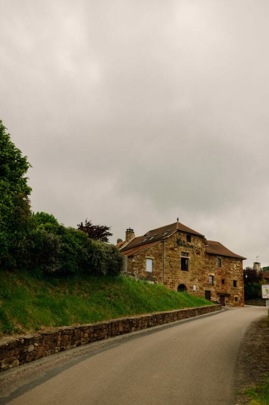 A stone house with a red-tiled roof sits beside a narrow, empty road under an overcast sky, surrounded by greenery and a stone wall. by Justin Salem Meyer