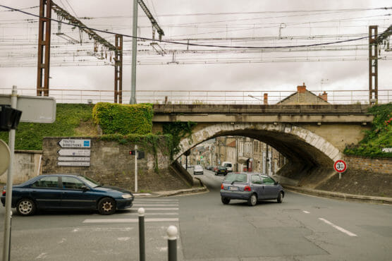 A few cars drive under an old stone railway bridge with overhead wires on a cloudy day in a small European town. Road signs and greenery are visible around the bridge. by Justin Salem Meyer