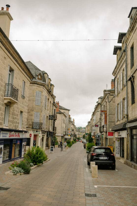A quiet, narrow street lined with stone buildings, shops, and balconies in a European town. Few people walk along the cobblestone path; a parked car is visible on the right. The sky is overcast. by Justin Salem Meyer