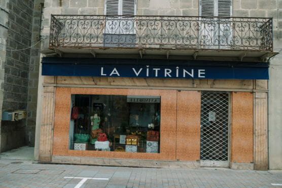 A storefront with a blue sign reading LA VITRINE above a large display window showing bags and clothing. The building has a balcony with a wrought iron railing and closed shutters above. by Justin Salem Meyer