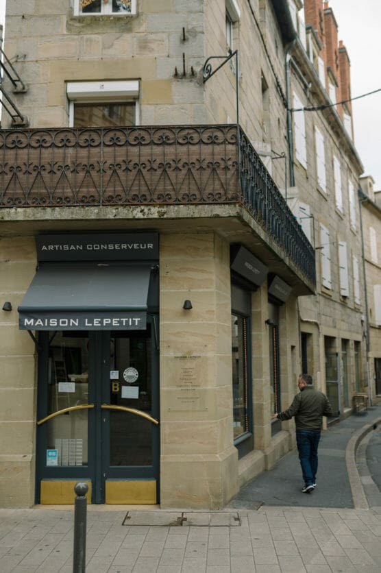 A man walks past the corner entrance of Maison Lepetit, a shop with stone walls, black awnings, and decorative iron balcony railings, on a narrow street in a European town. by Justin Salem Meyer