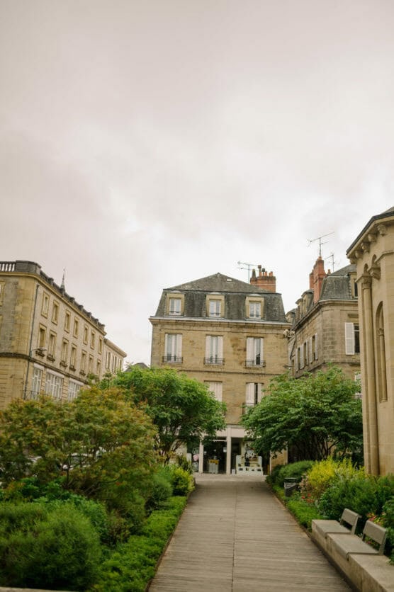 Pathway lined with green bushes and trees leads to a beige, three-story European building with white-trimmed windows and balconies, surrounded by other historic structures under an overcast sky. by Justin Salem Meyer