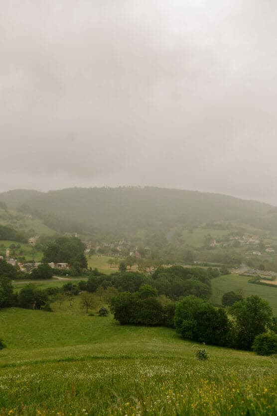 A misty landscape with rolling green hills, scattered trees, and small houses nestled in a quiet valley under a cloudy sky. by Justin Salem Meyer