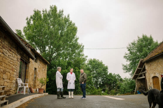 Three people stand talking in the courtyard of a rustic farm with stone buildings and trees in the background. Two wear white coats and boots, while the third wears casual clothes. A black animal is partially visible on the right. by Justin Salem Meyer