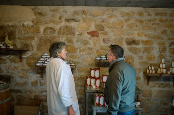 Two people stand in front of shelves with jars and cans against a rustic stone wall, one wearing a white coat and the other in a jacket, appearing to have a conversation. by Justin Salem Meyer