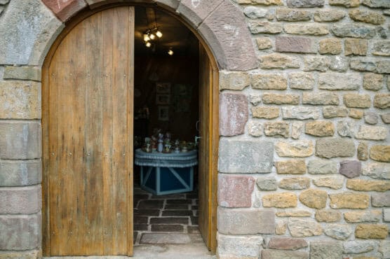 A wooden arched door partially open in a stone wall reveals a room inside with a round table displaying various small items, dimly lit by ceiling lights. by Justin Salem Meyer