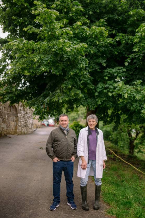Two people stand on a paved path beside a large green tree. One wears a jacket and scarf, the other a white coat and rubber boots. A stone wall and greenery are visible in the background. by Justin Salem Meyer