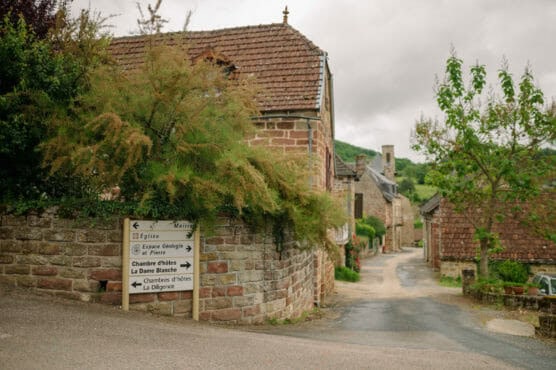 A narrow street in a quaint village with stone houses, greenery, and a road sign pointing to a church, guest rooms, and other village locations. The sky is overcast. by Justin Salem Meyer