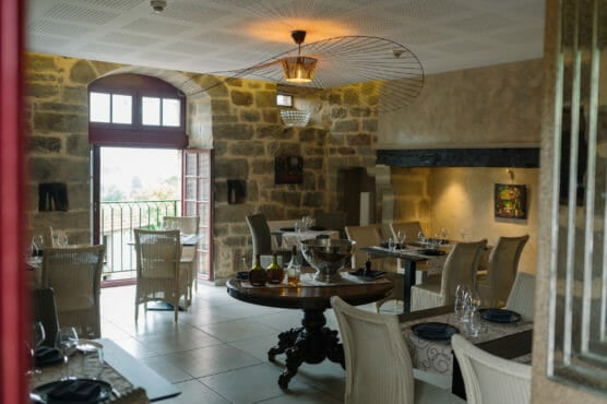 Elegant restaurant interior with stone walls, large windows, and a round central table set with wine glasses, a wine bucket, and candles. Wicker chairs and modern lighting create a cozy, inviting atmosphere. by Justin Salem Meyer