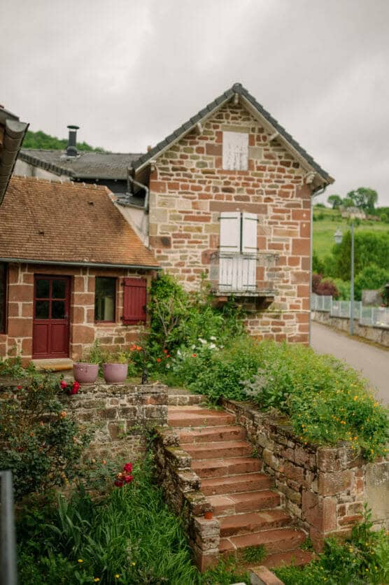 A rustic stone house with red shutters and white window, surrounded by lush greenery and flowers. Stone steps lead up to the entrance, and the sky is overcast. by Justin Salem Meyer