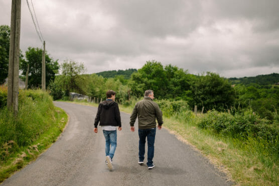 Two people walk side by side down a rural road surrounded by greenery and trees, under a cloudy sky. They both wear jackets and jeans, and their backs are to the camera. by Justin Salem Meyer