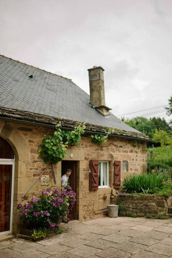 A stone cottage with a slate roof, brown wooden shutters, and a chimney sits in a garden. Green vines and purple flowers grow by the entrance, where a person peeks out of the door. The sky is overcast. by Justin Salem Meyer