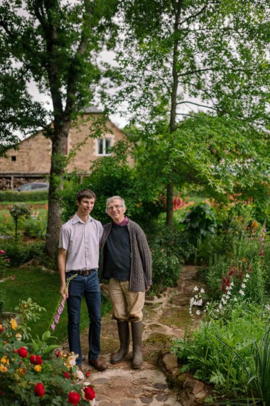 Two people stand on a stone path in a lush garden, surrounded by colorful flowers and green trees. One person wears a light shirt and jeans, holding a flower; the other wears a sweater, boots, and glasses. A house is visible in the background. by Justin Salem Meyer
