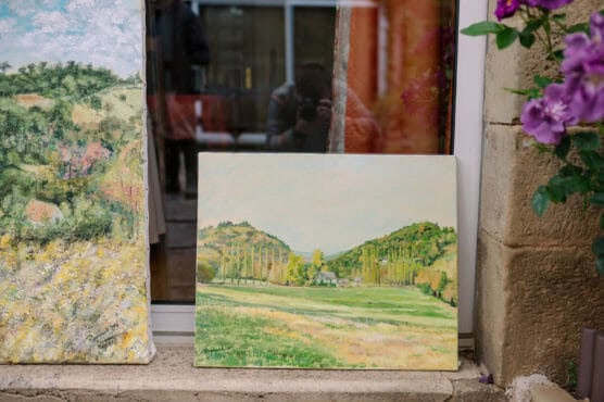 Two landscape paintings are propped outside against a window. One shows green hills and a field with trees, while the other is more abstract. Purple flowers grow nearby on the right. A faint reflection is seen in the glass. by Justin Salem Meyer