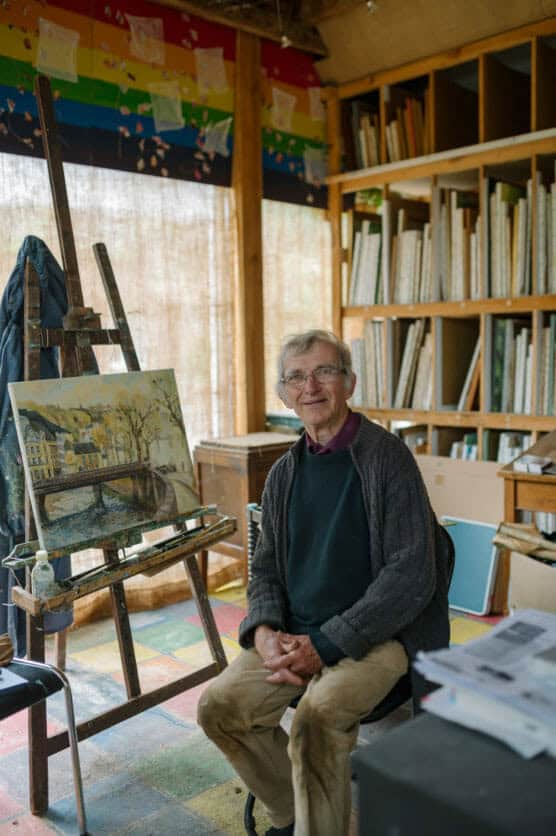 An older man with glasses sits smiling in an art studio, surrounded by bookshelves and paintings. An easel with a landscape painting is beside him. A colorful banner hangs above the window in the background. by Justin Salem Meyer