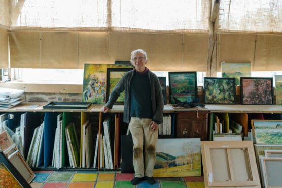 An older man with white hair stands in a colorful studio, surrounded by paintings on the walls and floor. He is smiling and resting one hand on a cabinet filled with framed artwork. Sunlight streams in through large windows covered with blinds. by Justin Salem Meyer