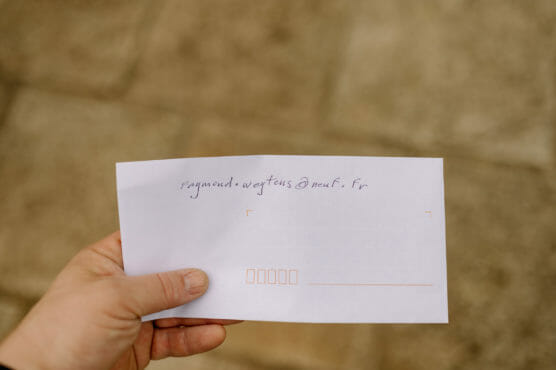 A hand holds a white envelope with handwritten text on it. The background is blurred stone or pavement. The envelope has some address lines and boxes printed in orange. by Justin Salem Meyer