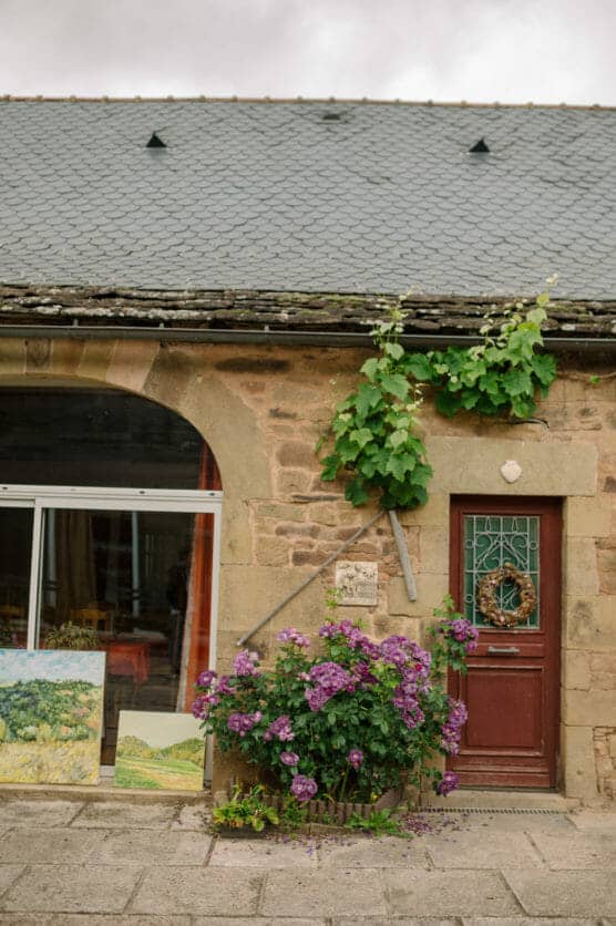 A rustic stone house with a red door, purple flowers, climbing vines, and paintings displayed outside near a large window, evoking a cozy, artistic atmosphere. by Justin Salem Meyer