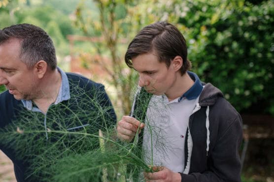 Two people stand outdoors among greenery. The younger person, wearing a gray hoodie, closely examines and smells a feathery plant, while the older person looks away, partially out of frame. Trees and shrubs are visible in the background. by Justin Salem Meyer