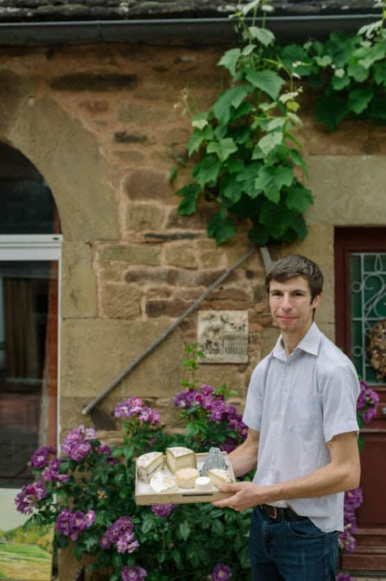 A man in a short-sleeve shirt stands outside a rustic stone building, holding a wooden tray with assorted cheeses. Purple flowers and green vines decorate the scene. by Justin Salem Meyer