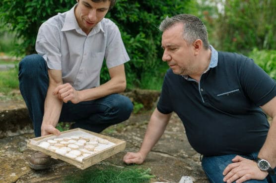 Two men kneel outdoors while examining a wooden tray filled with round cookies dusted with powdered sugar. One man holds the tray, and greenery is visible in the background. by Justin Salem Meyer