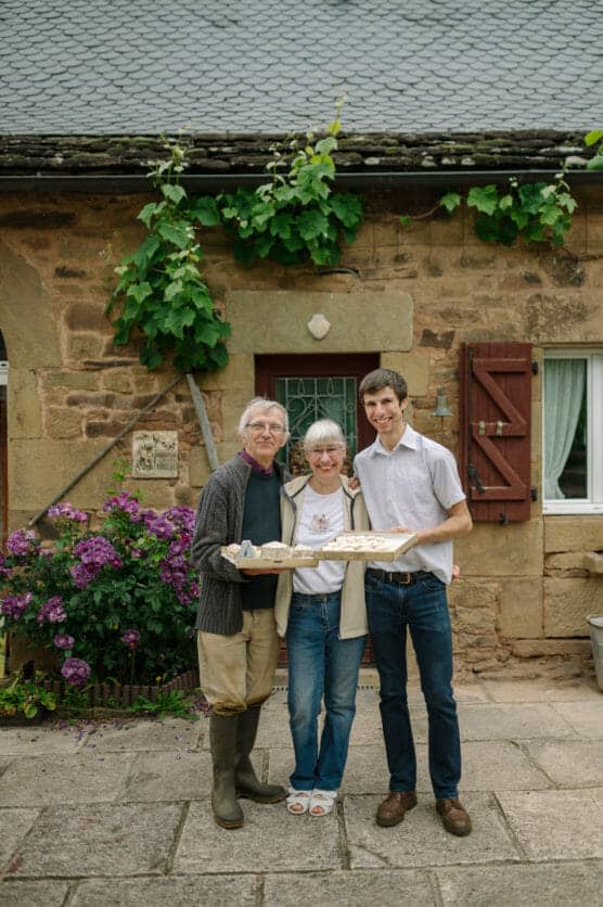 Three people stand smiling together outside a rustic stone house with green vines and purple flowers. The woman in the middle holds a tray of baked goods, and the two men stand on either side of her with arms around her. by Justin Salem Meyer