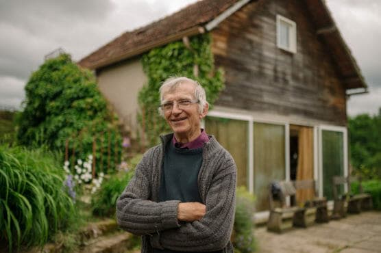 An older man with gray hair and glasses stands smiling with arms crossed in front of a rustic wooden house surrounded by lush greenery and plants. by Justin Salem Meyer
