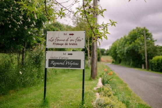 Two signs by a rural roadside point in different directions. One reads La Ferme den Fas, Fromage de brebis and the other Atelier d’artiste Raymond Wuytens. Lush greenery and a cloudy sky surround the area. by Justin Salem Meyer