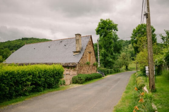 A narrow country road runs past a rustic stone house with a slate roof, surrounded by green bushes and trees under a cloudy sky. Power lines run along the roadside. by Justin Salem Meyer