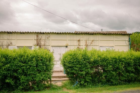 A single-story, cream-colored building with white doors and windows is partially obscured by a tall green hedge under a cloudy sky. by Justin Salem Meyer