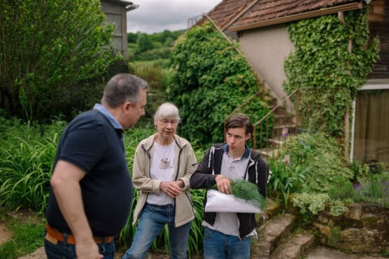 Three people stand outdoors by a house covered in ivy. One person holds a bag of green herbs, while the others talk to him. The setting is lush and green, with stone steps and plants in the background. by Justin Salem Meyer