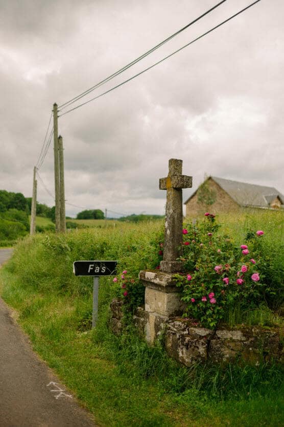 A stone cross stands beside a rural road, surrounded by blooming pink flowers and greenery. A signpost reads Fâts. Power lines and an old farmhouse are visible under a cloudy sky. by Justin Salem Meyer