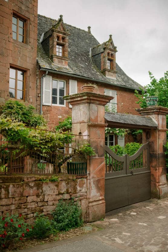 A charming old stone house with tall windows, a slate roof, and ornate gables stands behind a wrought iron gate and stone fence, surrounded by greenery and flowers on a cobblestone street. by Justin Salem Meyer