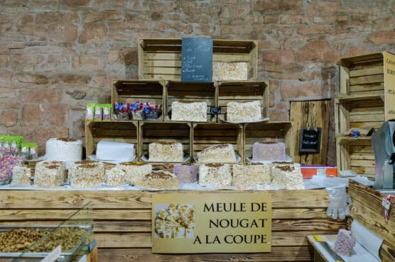 Various large blocks of nougat are displayed on wooden shelves and crates at a market stall, with a sign in French reading MEULE DE NOUGAT A LA COUPE and a brick wall in the background. by Justin Salem Meyer
