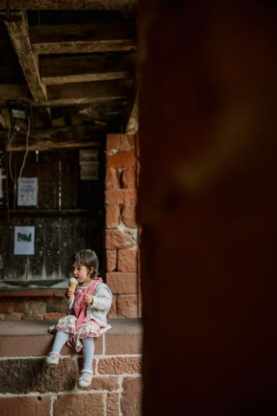 A young girl in a pink dress and white cardigan sits on a stone ledge, eating an ice cream cone. She is looking to the side, with rustic brick walls and a wooden roof in the background. by Justin Salem Meyer