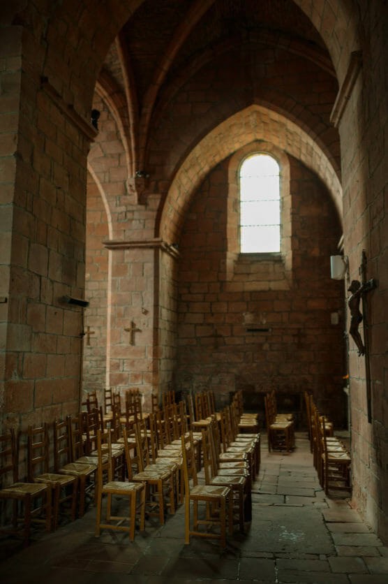 Rows of wooden chairs are arranged inside a dimly lit stone church with arched ceilings and a tall, narrow window letting in natural light. A crucifix is mounted on the right wall. by Justin Salem Meyer