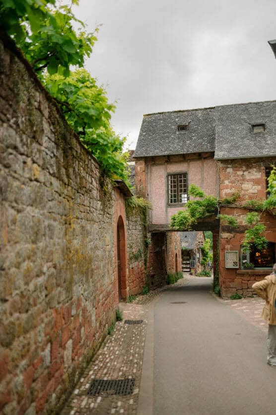 A narrow street in a quaint village with stone walls and a stone archway covered in greenery. An old building with a slate roof spans the street. A person stands partially visible on the right. The sky is overcast. by Justin Salem Meyer