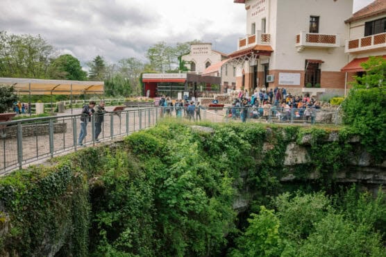 Visitors stand on a railing overlooking a lush, green chasm outside a building labeled Resurgence de la Sorgue. A group gathers near the entrance, surrounded by trees and gardens. by Justin Salem Meyer
