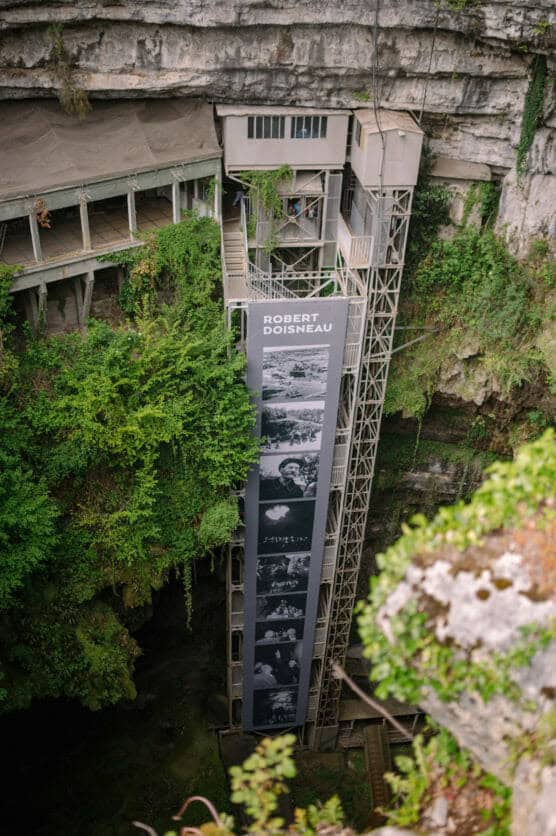 An outdoor elevator with photos and the name Robert Doisneau displayed on its side descends into a lush, green cave surrounded by rocks and vegetation. Elevated walkways are visible near the caves opening. by Justin Salem Meyer
