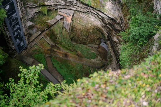 A deep, circular sinkhole with layered rock walls, featuring a series of wooden and metal walkways and stairs leading down into lush greenery at the bottom, surrounded by dense vegetation. by Justin Salem Meyer