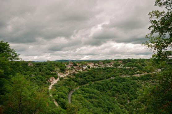 A small village with stone houses sits atop a lush, green hill surrounded by dense forest under a cloudy sky, with a winding river visible below. by Justin Salem Meyer